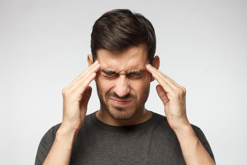 Portrait of young man isolated on gray background, suffering from severe headache, pressing fingers to temples with closed eyes | Revive MD Inc in Barrie, ON